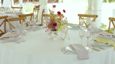 Şenlik masasında pembe renkli yabani çiçeklerden oluşan yakın plan bir buket var. Düğünde minimalist bir dekor, yuvarlak masa örtüsü, masada şarap bardakları ve tabaklar. Ağır çekim. 4k