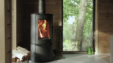 Küçük bir evde yanan şömine şöminesi, arka planda ahşap duvar ve yazın yeşil ormana bakan güzel pencere manzarası, yavaş çekim. Yüksek kalite 4k görüntü