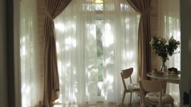 Mutfağın içi ahşap yuvarlak masa, üzerinde çiçekler olan vazo geniş pencerenin yanında dört ahşap sandalye ve sandalyedeki tüllerden güneş ışınları. Samimi iç tasarım. 4k 