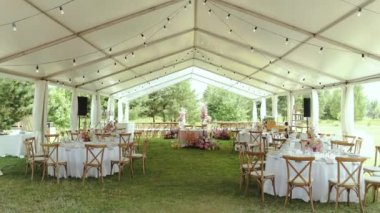 Çadırın altındaki düğün için geniş açılı dekore edilmiş masalar. Pembe renkli buketler şenlik masalarında, şarap bardaklarında ve tabaklarda, açık hava düğününde, ağır çekimde. 4k 
