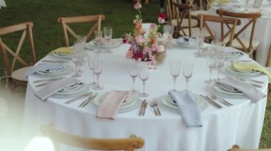 Şenlik masasında pembe renkli bir buket yabani çiçek var. Düğünde minimalist bir dekor, yuvarlak masa örtüsü, tahta sandalyeler, şarap kadehleri ve tabaklar. 4k