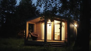 Gece evindeki ahşap İskandinavyalı pencerenin ışığı yanıyor, yazın teras sıcak elektrik ışığıyla yanıyor. Ormanın ortasında sessiz bir orman kulübesi, gece gökyüzü, ağır çekim. 4k görüntü