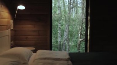 Close-up of cozy scandinavian illuminated bedroom with a bedside lamp and white double bed covered by grey sheet cover, wooden wall and view from the window to the forest, slow motion. 4k footage