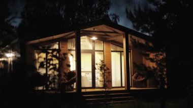 Ahşap İskandinav evinin gece çekimleri pencerenin ışığı açık, yazın sıcak elektrik ışığıyla sıcak teras. Ormanın ortasında sessiz bir orman kulübesi, gece gökyüzü, ağır çekim. 4k 
