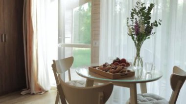 Bir vazo çiçekle süslenmiş ahşap bir yemek masası pencerenin kenarında oturur, doğanın dokunuşuyla evin iç tasarımını güçlendirir.