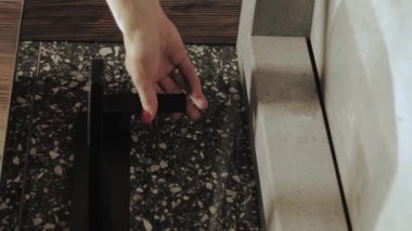 Bir kadının eli banyoda siyah bir musluğu çevirir. Su akıyor. Karanlık banyo, siyah musluk ve mermer lavabo, yavaş çekim. Yüksek kalite 4K görüntü. Dikey video