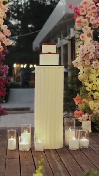 結婚式のアーチの背景に対するウェディングケーキは 花とろうそくで装飾され 魔法の雰囲気を醸し出し 部屋に自然のタッチを追加します バーティカルビデオ — ストック動画