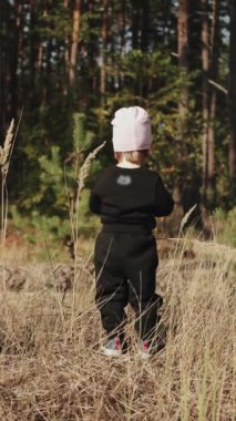 Dikey video. Genç bir çocuk, güzelliklerle dolu çarpıcı bir orman zeminiyle çevrili, uzun, yemyeşil çimlerin üzerinde neşeyle duruyor.