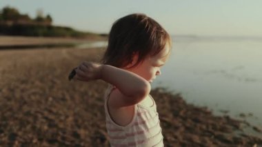 Güneş durgun suyun üzerinde yavaşça batarken küçük bir çocuk elinde bir sopayla kumsalda neşeyle geziyor.