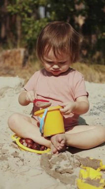 Neşeli bir çocuk kumlu bir sahilde renkli oyuncaklarla yaratıcı oyunlarla meşgul, sevimli doğayla çevrili. Dikey video