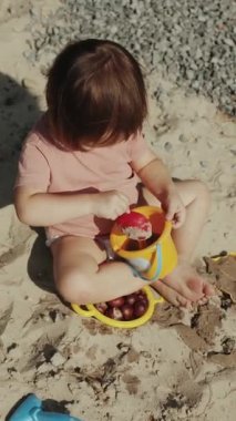 Genç bir çocuk neşeli bir şekilde parlak, güneşli bir sahil bölgesinde canlı ve renkli kum oyuncaklarıyla oynamaktan zevk alır. Dikey video. 