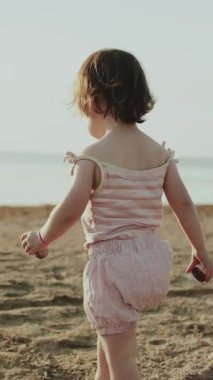 Neşeli bir çocuk neşeli bir şekilde kumlu kumsalları keşfeder, canlı bir günbatımının sıcaklığını kucaklayarak. Dikey video. 