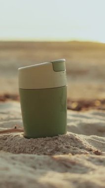 Dikey video. Yeniden kullanılabilen şık bir kupa kumlu bir plajda oturur, eko-arkadaşça titreşimler ve açık havada rahat bir yaşam tarzı.