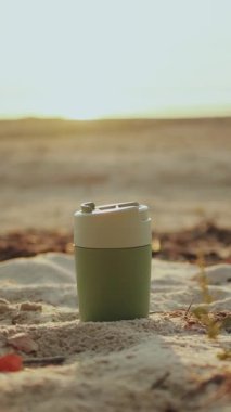 Sıcak kumun üzerindeki modern bir seyahat kupası yaz akşamları gün batımında plaj sükunetinin özünü yakalar. Dikey video