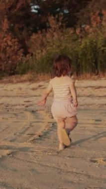 Dikey video. Genç bir çocuk neşeyle kumlu plajı keşfediyor, nefes kesici günbatımının güzel renklerine dalmış.