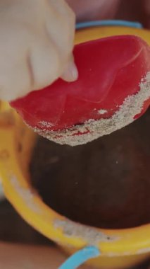 Dikey video. Bir çocuk, yanında parlak sarı bir kova ile kumu kazarken kırmızı bir kürekle mutlu bir şekilde oynuyor.