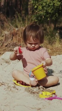 Dikey video. Genç bir çocuk büyüleyici doğal ortamında çeşitli renkli oyuncaklarla sıcak kumlu sahillerde neşeyle oynuyor.