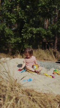 Dikey video. Neşeli genç bir çocuk bereketli ağaçlarla çevrili kumlu bir alanda çeşitli renkli oyuncaklarla enerjik bir şekilde oynar.