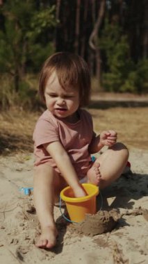 Neşeli bir çocuk, sıcak kumda parlak turuncu bir kovayla güzel bir doğal ortamda neşeyle oynar. Dikey video. 