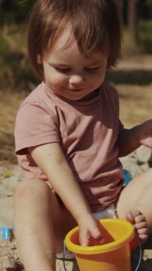 Dikey video. Neşeli ve neşeli bir çocuk, canlı bir güzelliğe dalmış, parlak sarı bir kovayla hevesle meşgul oluyor.