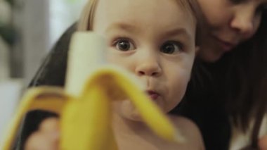 Neşeli ve mutlu bir çocuk, bir bakıcı tarafından sevgiyle desteklenirken lezzetli bir muzun içinde keyiflenir.