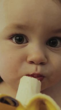 Dikey video. Neşeli bir çocuk heyecanlı bir şekilde lezzetli bir muzu tıkınır, neşeli ve kaygısız yemek anından zevk alır.