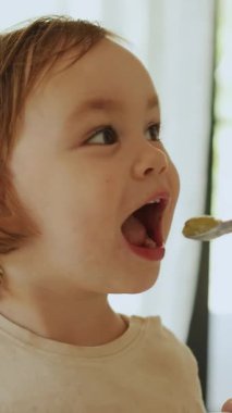 Çocuk yemeğin ve gülümsemenin tadını çıkarıyor, anne masada kaşıkla çocuğu besliyor. Dikey video