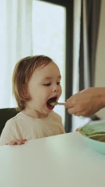 Dikey video. Neşeli bir bebek yemekten zevk alır, bir anne çocuğu masada kaşıkla besler. Sıcak anılar oluşturuluyor.