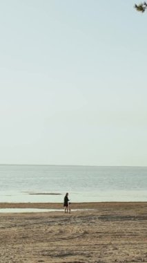 Dikey ekran: Huzurlu bir sahil boyunca yürüyen yalnız bir figür, engin, mavi bir gökyüzü altında, sükunet ve yalnızlık hissi uyandırıyor