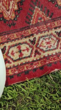 Dikey ekran: zarif ve canlı geleneksel bir halı, verimli çimenli bir arka plana karşı ustalıkla karışık desenler sergiliyor.