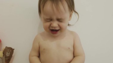 Yürümeye başlayan bir çocuk beyaz bir duvarın önünde neşeli ve neşeli bir şekilde dururken güler. Giysileri olmayan bir çocuk, güzel küçük bir kız. 4k