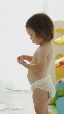 Neşeli bir çocuk, parlak, güneşli bir odada bebek bezi giyip, çevresini keşfediyor. Dikey video 4k