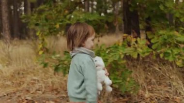 Mutlu ve gülümseyen bir çocuk elinde peluş bir köpek, sıcak sonbahar ormanlarında koşar ve geri döner. Yüksek kalite 4k görüntü