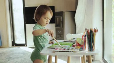 Konforlu bir evde, küçük bir masada parlak renkler çizen bir çocuk, küçük bir evin parlak bir odasının penceresinden dışarı bakıyor. Yüksek kalite 4k görüntü