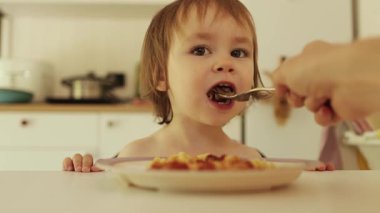 Bir bebek evdeki rahat ve parlak beyaz bir mutfakta bir kase yulaf lapası yiyebilir. Bir anne çocuğunu kasede besler. Yüksek kalite 4k görüntü