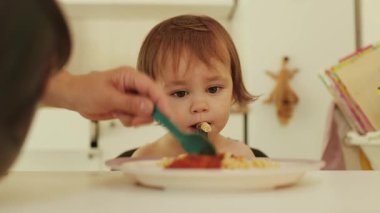 Parlak bir mutfakta beyaz bir masada oturan çocuk makarna yiyor. Sevimli ve keyifli bebek baharatlı soslu renkli bir tabak spagetti bekliyor. Yüksek kalite 4k