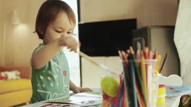Bir bebek, parlak bir odada beyaz bir masanın üzerine fırçayla resim çizerek sanatsal yönünü yaratıcı bir şekilde renkli boyalar ve çeşitli fırçalar kullanarak evinde keşfeder. Yüksek kalite 4k görüntü