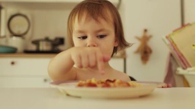 Üç yaşında bir kız yemek tabağını işaret ediyor. Meraklı çocuk parmaklarıyla lezzetli makarnayı keşfediyor, tat, doku ve gülümsemeyi hissediyor. Yüksek kalite 4k görüntü