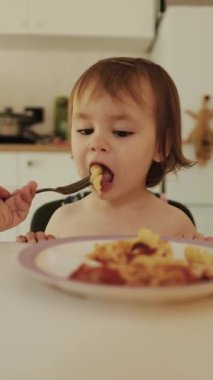 Sıcak bir mutfakta bulunan neşeli ve neşeli bir bebek çatalla lezzetli ve renkli bir makarna ile beslenir.