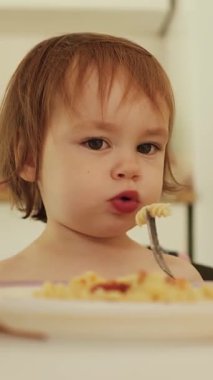 Dikey çekim. Bir kız masada otururken sıcak makarna üflüyor. Annem çocuk için yiyecek dolduruyor. Çocuk evde rahat bir şekilde makarnanın tadını çıkarıyor.