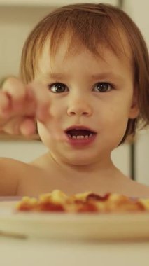 Dikey çekim. 3 yaşında bir kız masaya uzanıp makarnayı parmağıyla yiyeceği işaret ediyor. Yüksek kalite 4k görüntü