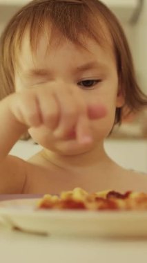 Dikey video. Kız yemek tabağını işaret ediyor. Yeni yürümeye başlayan çocuk lezzetli makarnayı parmak, tat ve dokularla keşfediyor ve gülümsüyor. Yüksek kalite 4k görüntü