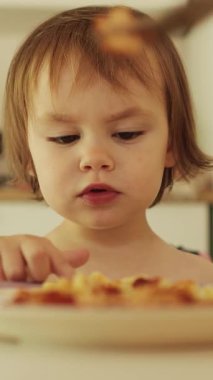 Dikey çekim. Çocuk, annesinin tabağın üzerindeki kaseden ona verdiği yemeği yemek istemiyor. Parmağıyla başka bir şey istediğini işaret ediyor. Kız makarna yiyor. Yüksek kalite