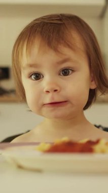 Dikey video. Neşeli ve mutlu 3 yaşındaki çocuk, sıcak ve parlak bir mutfakta domates soslu spagetti yemekten hoşlanıyor. Yüksek kalite 4k görüntü
