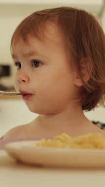 Dikey çekim. Çatallı bir anne, parlak beyaz bir mutfakta üç yaşındaki çocuğunu besler. Küçük kız makarna yiyor. Küçük bebek mutlu bir şekilde sıcak bir mutfakta lezzetli yemekler yiyor.