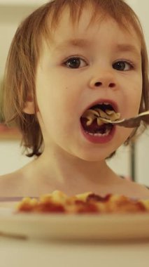 Dikey video. Yeni yürümeye başlayan çocuk, evdeki rahat ve parlak beyaz bir mutfakta makarna yiyor. Anne çocuğunu bir kaseden besliyor. Mutlu çocuk. Yüksek kalite 4k görüntü