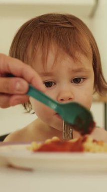 Dikey video. Parlak bir mutfakta açık bir masada oturan çocuk makarna yiyor. Anne yemek koyuyor ve bebek hevesle baharatlı soslu renkli bir tabak spagetti bekliyor. Yüksek kalite 4k