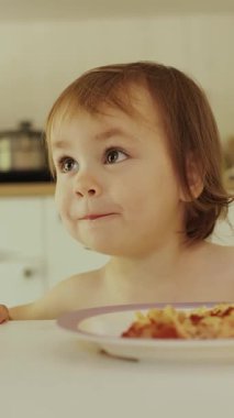 Dikey video. Anne, parlak mutfaktaki masada mutlu çocuk makarnasıyla besleniyor. Bebecik lezzetli yemeklerden zevk alıyor, aile bağlarının önemini vurguluyor. Yüksek kalite 4k görüntü