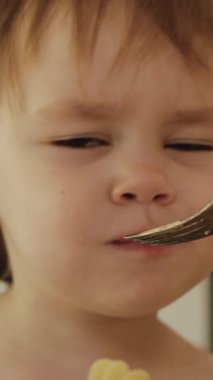 Dikey video. Mutlu çocuk portresi. Parlak mutfakta lezzetli yemekler yiyor, el sallıyor ve gülümsüyor, makarna yiyor. Anne, çocuğunu masada besliyor. Yüksek kalite 4k görüntü