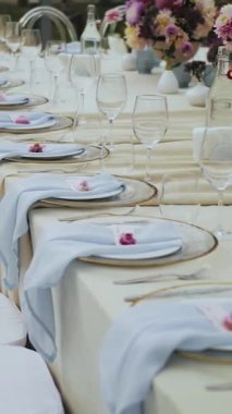 Dikey video. Düğün ya da etkinlikte pembe çiçeklerle süslenmiş, tabaklar ve bardaklarla süslenmiş bir düğün masası. Şenlik masasının yanındaki beyaz sandalyeler. Çiçek düzenleme masada, yavaş çekim.